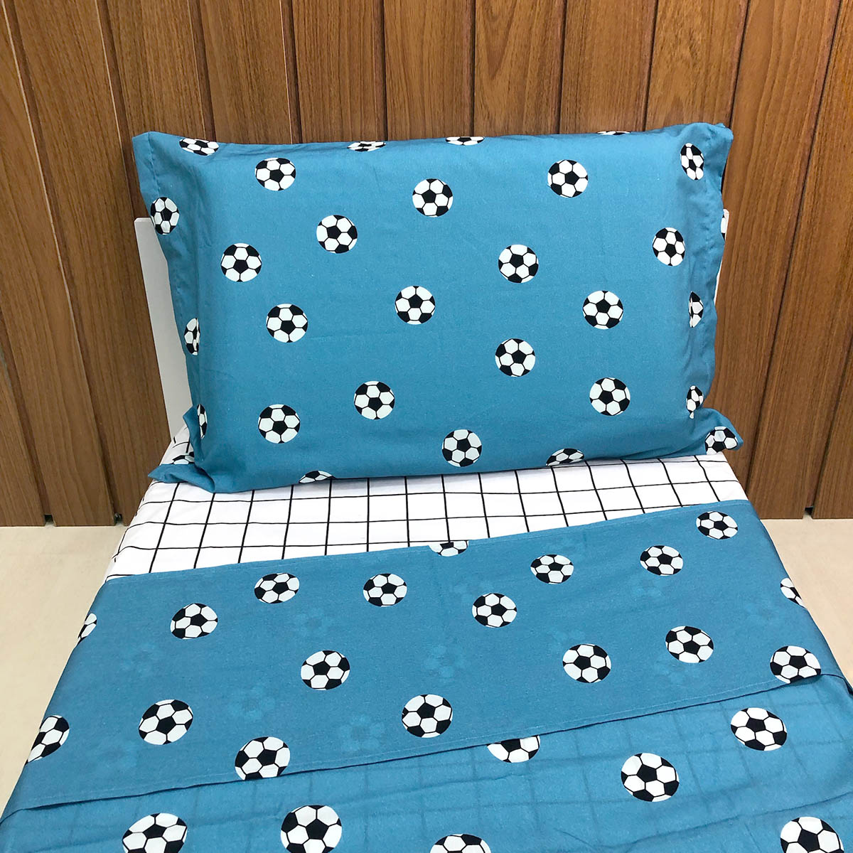 Jogo de cama infantil com bolas de futebol com fundo azul na fronha e lençol de cobrir. O lençol de elástico é quadriculado preto e branco.
